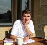 A photo of Joe Redmayne, a young man with brown hair in a white s-shirt. He is wearing a white t-shirt and writing in a notebook, looking into the camera.