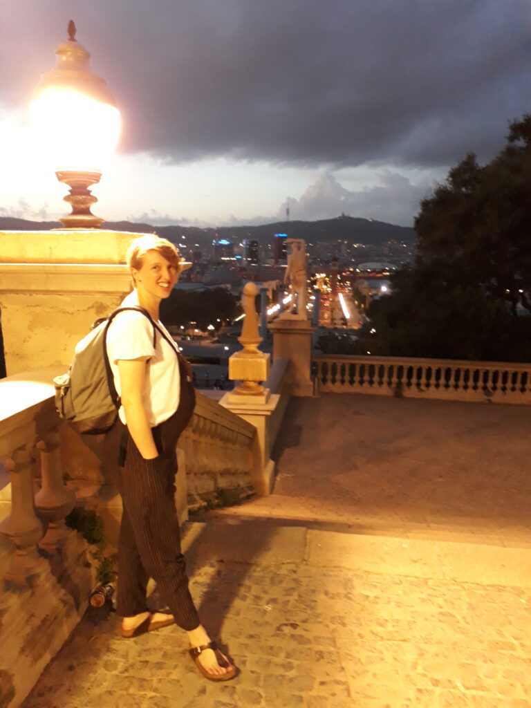 A heavily pregnant person smiling at the camera on a set of pretty stairs, looking out at the Barcelona skyline.