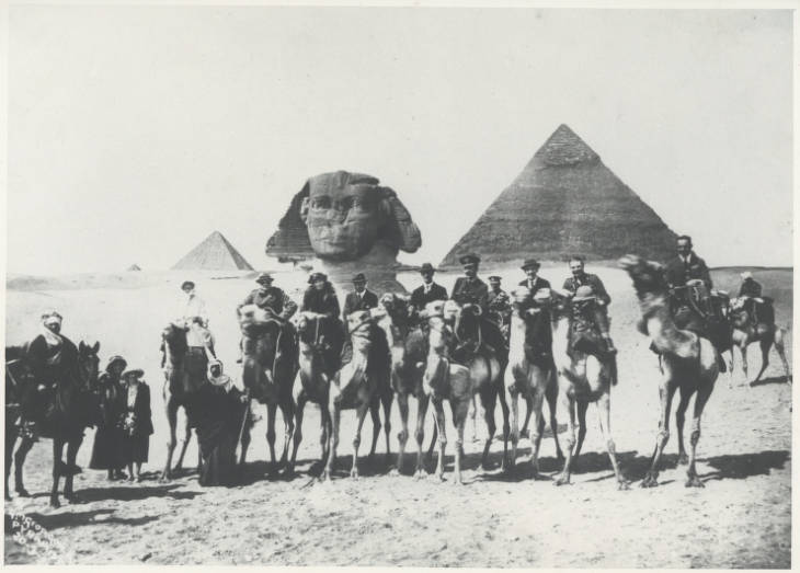 Photograph of a group of riders on camels with the Pyramids in the background.
