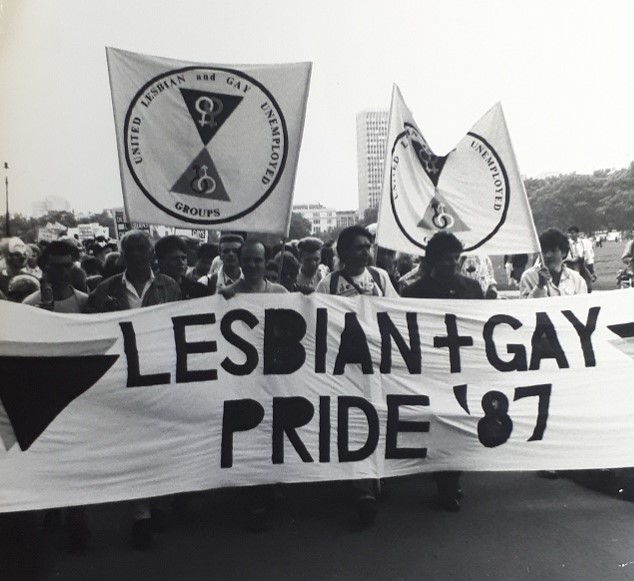 Photograph of London Pride 1987 showing a group of people carrying a banner with 'LESBIAN + GAY PRIDE '87' written in bold letters on it.