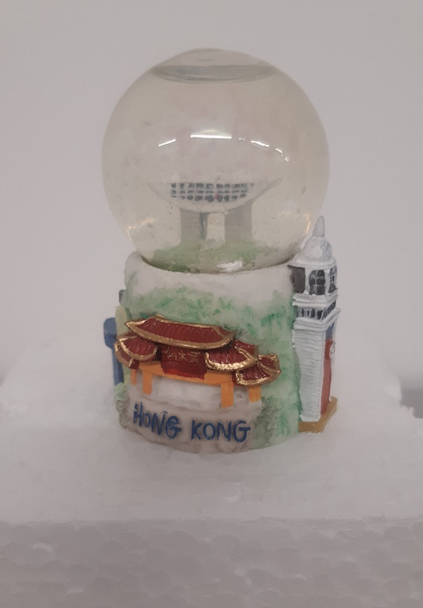 A small snow globe on a polystyrene base. The snow globe has a white, green and red base with the term 'hong kong' along the base and ornamental towers along the side of the base. Inside the snow globe is a small scale feature of the Peak tower, consisting of 2 columns supporting a  wide dish building element. 