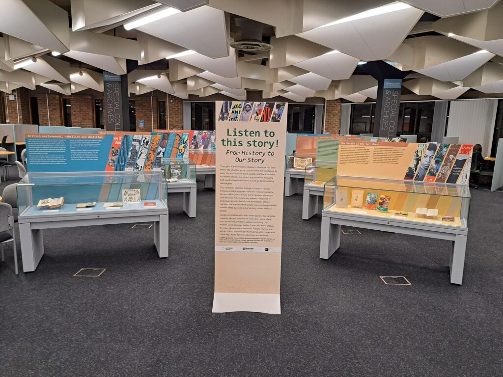Listen to this story! exhibition in situ, showing a tall banner with text in the middle and 6 staggered exhibition cases behind each other to the left and right of the banner.