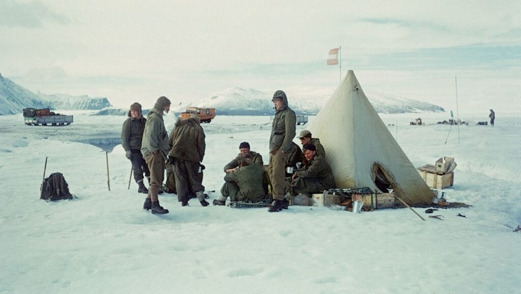 Coloured photograph showing members of the British North Greenland Expedition in 1952, sitting and standing outside a tent on a glacier in Greenland