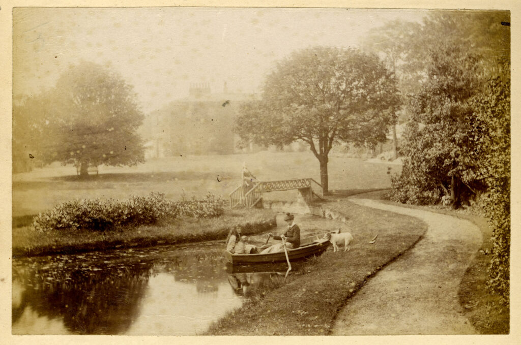 Sepia photograph of the a person in a both with their door at the side, boating on the fishpond, with the house in the background